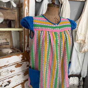 May include: A vintage-style, colorful tunic dress with a striped pattern in pink, green, and yellow. The dress features blue accents on the sleeves and pockets. The garment is displayed on a mannequin.