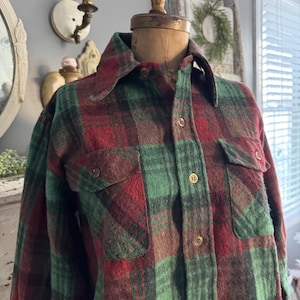Puede incluir: Una camisa de franela vintage con un patrón de cuadros rojos, verdes y marrones. La camisa tiene cuello y dos bolsillos en el pecho. La tela parece gruesa y cálida. La camisa es abotonada y parece hecha para climas más fríos.