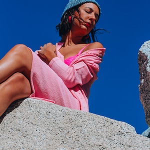 Peut inclure: Une femme portant un cardigan rose et un soutien-gorge rose est assise sur un rocher avec un ciel bleu en arri&egrave;re-plan. Elle porte un bonnet bleu et des boucles d'oreilles rouges.