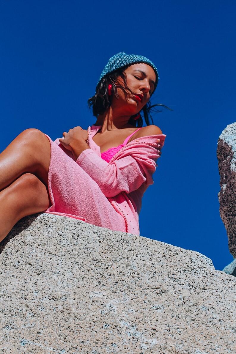 Peut inclure: Une femme portant un cardigan rose et un soutien-gorge rose est assise sur un rocher avec un ciel bleu en arri&egrave;re-plan. Elle porte un bonnet bleu et des boucles d'oreilles rouges.