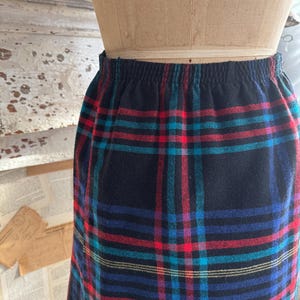 May include: A vintage, knee-length skirt with a classic plaid pattern. The skirt features a black base with a colorful plaid design in red, blue, teal, and yellow. The waistband is elasticized. The skirt is displayed on a mannequin.