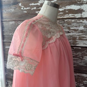Op de afbeelding: Een vintage roze nachtjapon op een mannequin. De japon heeft een doorschijnende stof met kanten afwerking rond de halslijn en mouwen. De korte mouwen zijn versierd met kleine roze strikjes. De nachtjapon heeft een vloeiend, geplooid ontwerp.