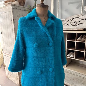 May include: A vintage turquoise double-breasted coat with a textured, plush appearance. The coat features a classic collar, three-quarter sleeves, and large, round buttons. The coat is displayed on a vintage mannequin.