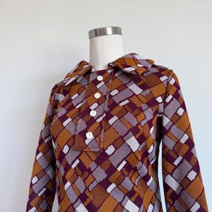 May include: A long-sleeved dress featuring a geometric pattern in shades of mustard yellow, deep burgundy, and light gray. The dress has a wide collar and a front placket with four white buttons, displayed on a mannequin torso.