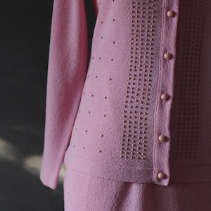 Peut inclure: Un ensemble jupe et cardigan en tricot rose avec un motif perl&eacute; et des boutons.
