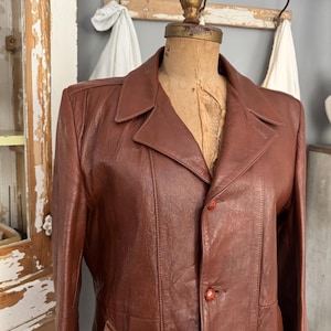 Puede incluir: Chaqueta de cuero marrón vintage con cuello de solapa clásico y tres botones rojos. La chaqueta se exhibe en un maniquí, mostrando su ajuste a medida y su rico color. La chaqueta tiene un bolsillo en el lado izquierdo.