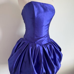 May include: A strapless, vibrant royal blue dress featuring a fitted bodice and a voluminous, pleated bubble skirt. The garment is crafted from a smooth, satin-like fabric that reflects light, creating a structured silhouette on a display mannequin.