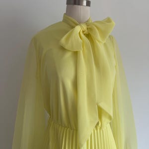 Puede incluir: Vestido amarillo pálido con cuello alto y lazo grande. El vestido presenta mangas largas y transparentes y una falda plisada. El vestido se exhibe en un maniquí sobre un fondo blanco liso.