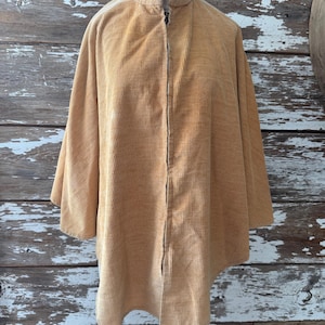 May include: A tan corduroy jacket with a high collar and a full-length zipper. The jacket has a relaxed fit and three-quarter length sleeves. The fabric has a ribbed texture. The jacket is displayed on a mannequin against a weathered wood background.