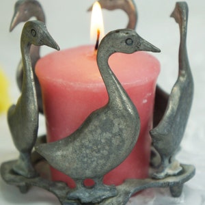 May include: A silver candle holder shaped like four geese surrounding a pink candle.