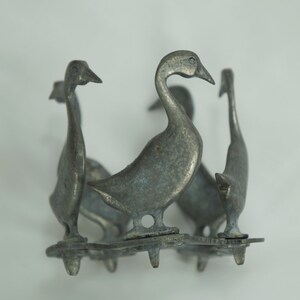 May include: A silver metal goose figurine with four geese standing in a circle. The geese are facing outward and have a textured surface.