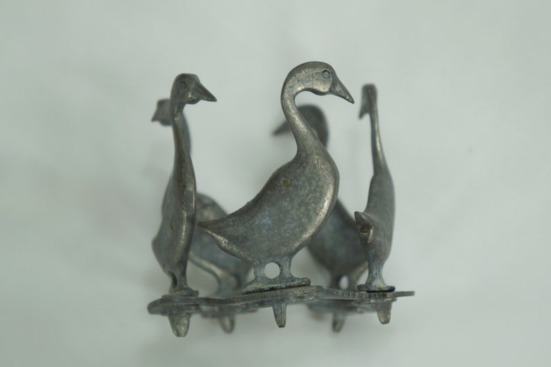 May include: A silver metal goose figurine with four geese standing in a circle. The geese are facing outward and have a textured surface.
