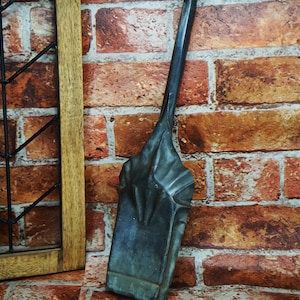 May include: A black metal fireplace shovel with a long handle. The shovel is leaning against a brick wall.