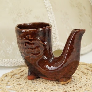 May include: A brown ceramic boot-shaped shot glass with a decorative design. The glass is sitting on a white crocheted doily.