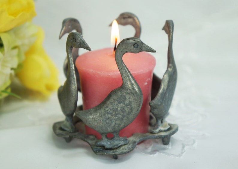 May include: A silver candle holder shaped like four geese surrounding a pink candle.