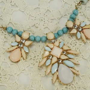 May include: A statement necklace with a large, multi-faceted pendant featuring pastel blue, pink, and white stones. The pendant is surrounded by smaller stones and is attached to a turquoise beaded chain.