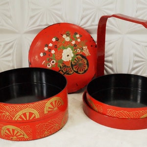 Puede incluir: Una caja bento japonesa de dos niveles roja y dorada con asa. El nivel superior tiene un diseño floral y el nivel inferior un patrón de ondas.