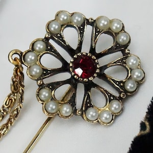 Puede incluir: Un broche dorado con un centro de pedrería roja y un delicado diseño floral. El broche está adornado con perlas falsas y tiene una cadena adjunta.