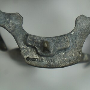 May include: Close-up of a silver-toned metal object with a detailed design. The object has a pointed tip and a circular inscription that reads "METZKE 170A".