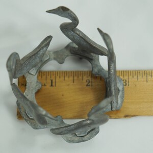 May include: A silver metal birdcage shaped like a circle with six birds, each with a long neck and beak, forming the cage. The cage is about 2 inches in diameter and is sitting on a wooden ruler.