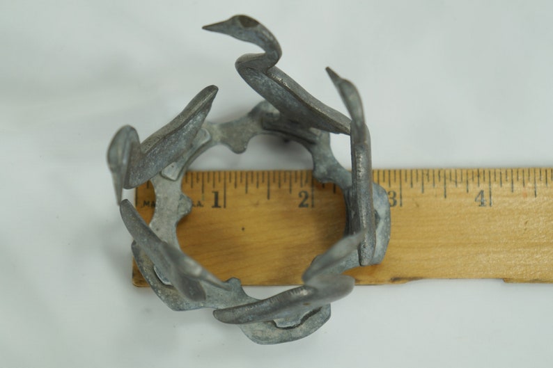 May include: A silver metal birdcage shaped like a circle with six birds, each with a long neck and beak, forming the cage. The cage is about 2 inches in diameter and is sitting on a wooden ruler.