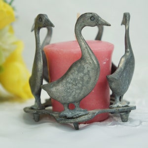 May include: A silver candle holder shaped like four geese surrounding a pink candle.