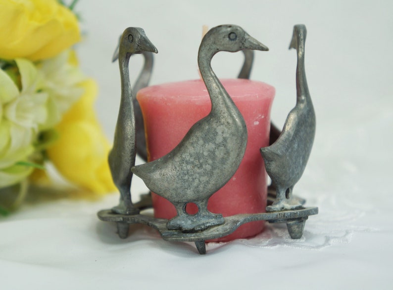 May include: A silver candle holder shaped like four geese surrounding a pink candle.