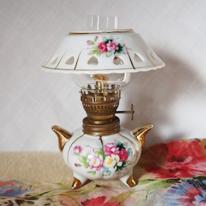 May include: A white porcelain oil lamp with a floral design and a gold trim. The lamp has a clear glass chimney and a white porcelain shade with a cut-out design.