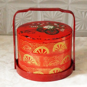 Puede incluir: Una fiambrera roja y dorada de varios niveles con asa. El nivel superior tiene un diseño floral, mientras que los dos niveles inferiores tienen un diseño de olas y sol.