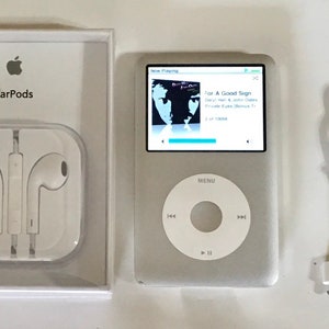 May include: A silver digital music player with a white circular click wheel and a color screen displaying album art, accompanied by white wired EarPods in their original packaging and a white 30-pin charging cable tied with a blue rubber band.
