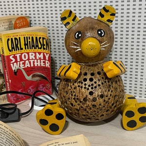 Pode incluir: Uma lâmpada de coco em forma de tigre com listras amarelas e pretas. A lâmpada tem um cabo e um interruptor. A lâmpada está sobre uma mesa com um livro.