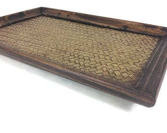 Può includere: Un vassoio rettangolare in legno con una base in bambù intrecciato. Il vassoio ha una finitura marrone scuro ed è perfetto per servire cibo o bevande.