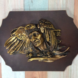 Puede incluir: Una escultura de pared dorada que representa a un hombre nativo americano con un águila posada en su hombro. La escultura está montada en una placa de madera marrón oscuro.