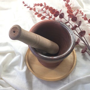 May include: A brown ceramic mortar and pestle set on a wooden base. The mortar is round and has a rough texture. The pestle is made of wood and has a smooth surface. The set is on a white background.