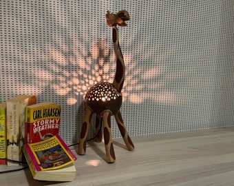 Op de afbeelding: Een houten giraffenlamp met een kokosnootschelplichaam en een licht erin. De lamp staat op een houten oppervlak met een stapel boeken ervoor.