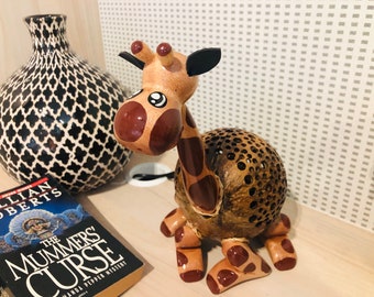 Op de afbeelding: Een houten giraffenbeeldje met bruine en witte vlekken. De giraffe is gemaakt van een kokosnootschelp en heeft een lichtbruin lichaam met bruine vlekken. De giraffe heeft een zwart oor en een bruin gezicht met witte ogen.