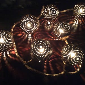 Może przedstawiać: Girlanda świetlna wykonana z kokosów z misternymi rzeźbionymi wzorami. Każdy kokos jest podświetlony od wewnątrz, dając ciepłe światło.