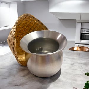 Może przedstawiać: Szlifowany metalowy garnek z szerokim, rozkloszowanym brzegiem i zaokrąglonym korpusem. Za garnkiem znajduje się tkany kosz z jasnobrązowego bambusa. Przedmioty znajdują się na szarym blacie w kuchni.