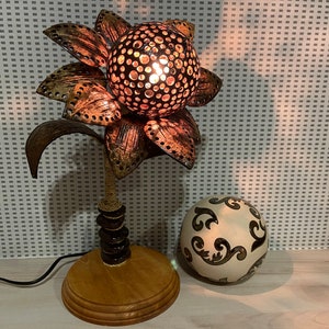 Pode incluir: Um abajur de mesa feito de uma casca de coco com um design de flor. A lâmpada está acesa e tem uma base de madeira marrom. Uma esfera branca decorativa com um design preto está no fundo.