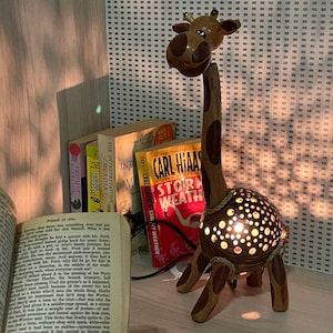 May include: A wooden giraffe-shaped lamp with a light shining through the body. The lamp has a brown and tan color scheme with a rope accent. Several books are in the background, including one open book.