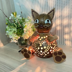 May include: A brown coconut shell cat lamp with a light brown face and white eyes. The lamp has a cutout design and is lit from within, casting a warm glow. The cat is sitting on a wooden surface.
