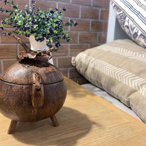 Puede incluir: Una caja de madera marrón oscuro con una tapa con bisagras y tres patas. La caja está hecha de un material natural, posiblemente cáscara de coco. La caja está sentada sobre una mesa de madera.