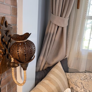 May include: A wooden wall sconce with a carved design holds a coconut shell lampshade. The lampshade has intricate cutouts and is lit by a small light bulb. The sconce is mounted on a brick wall next to a window with white curtains.