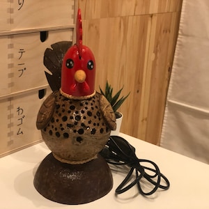 Puede incluir: Una lámpara de coco marrón en forma de gallo con cabeza roja y cable negro. La lámpara está sobre una mesa blanca.