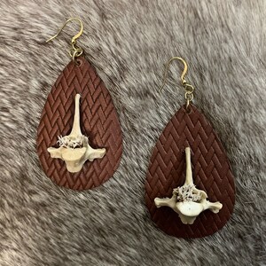 Op de afbeelding: Een paar bruine leren oorbellen in druppelvorm met een witte, bot-achtige hanger in het midden. De oorbellen hebben een geweven patroon.