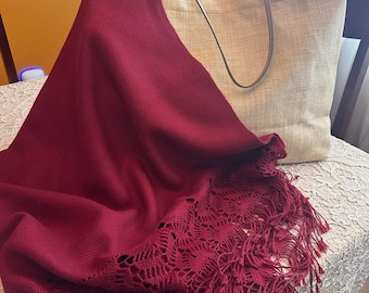 May include: A burgundy shawl with a decorative, openwork border and fringe is draped over a beige tote bag. The bag has a simple design with long, thin handles. The items are on a lace-covered surface.