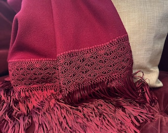 May include: A burgundy-colored throw blanket with a decorative patterned border and long fringe. The blanket is draped over a beige tote bag, creating a layered effect. The fringe adds texture and visual interest to the design.