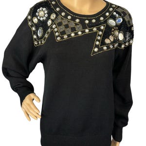 May include: Black sweater with embellished shoulders. The sweater has a crew neck and long sleeves. The shoulders are decorated with sequins, beads, and rhinestones in gold, silver, and clear colours. The sweater is a solid black colour.
