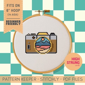 Könnte beinhalten: Ein Kreuzstichmuster in einem Holzrahmen mit einer braunen Kamera und einem kreisförmigen Berglandschaftsmotiv. Der Text lautet: FITS ON 6" HOOP (14 AIDA) BEGINNER FRIENDLY. Weitere Texte sind HIGH STRUNG cards, PATTERN KEEPER, STITCHLY und PDF FILES.