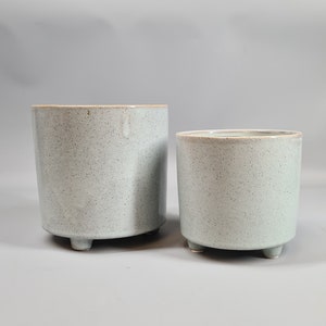 May include: Two grey ceramic planters with a speckled finish and three small feet on the bottom. The larger planter is in the background and the smaller planter is in the foreground.
